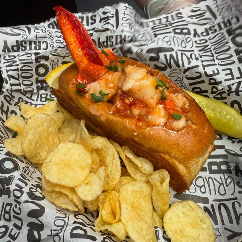 The Hungry Captain's lobster roll with a claw, chips, lemon, and pickle on branded paper.