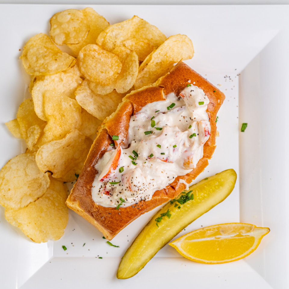 The Hungry Captain's lobster roll with creamy filling, chips, a pickle spear, and lemon wedge on a white plate.
